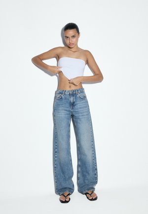 Jeune femme ajustant son top tube blanc, portant un jean bleu ample et des tongs noires, debout devant un fond blanc uni.