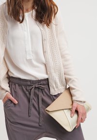 Beige gebreide cardigan met een textuurpatroon, witte blouse, grijze shorts met trekkoord en een envelop clutch met gouden accenten.