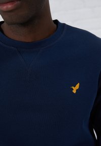 Marinblå sweatshirt i mjukt tyg med en gul broderad kolibri-logga på bröstet och en klassisk rund halsringning.