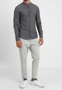 Chemise grise à manches longues avec col mandarin, boutons sur le devant, portée avec un pantalon beige clair fuselé et des baskets blanches.