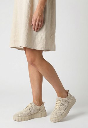 Kvinde iført beige hæklet sneakers og en matchende beige kjole, stående mod en ensfarvet lys baggrund.