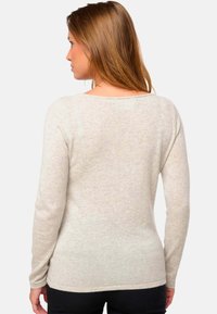 Femme aux longs cheveux châtains clairs portant un pull à manches longues beige clair, de dos contre un fond blanc uni.