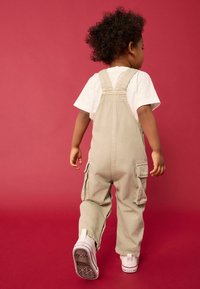 Salopette en coton beige avec bretelles et poches latérales, portée par-dessus un t-shirt blanc, avec des baskets blanches sur fond rouge.