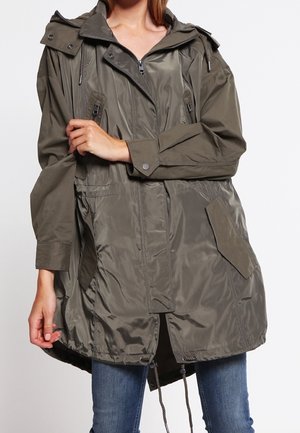 Femme portant une veste anorak à capuche vert olive avec fermeture éclair devant, boutons-pression, poches à rabat et cordons, associée à un jean bleu.