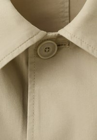 Gros plan du col de chemise beige avec un bouton rond fixé à travers la boutonnière.