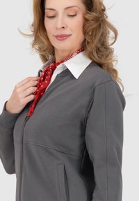 Donna con capelli ondulati che indossa una giacca zip grigia, una camicia bianca con colletto e una sciarpa rossa a pois, che guarda in basso mentre tocca la sciarpa.