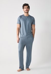 Young man standing barefoot, wearing a blue t-shirt and matching pants, with a neutral background. He looks directly at the camera.
