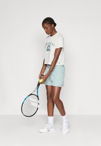 Világosszürke, zöld mintával ellátott, rövid ujjú póló, mentazöld sorttal párosítva. Teniszütőt és labdát tart, fehér sportcipőt és zoknit visel.