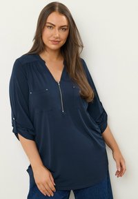 Marineblaue Bluse mit einem Reißverschluss vorne, zwei Vorderaschen und hochgekrempelten Ärmeln. Aus glattem Stoff mit einer lockeren Passform und abgerundetem Saum.
