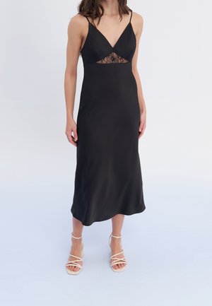 Vrouw draagt een zwarte mouwloze midi-jurk met kantdetail onder de buste en witte sandalen met hoge hakken en bandjes, staande op een effen achtergrond.