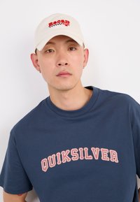 Νεαρός άνδρας με καπέλο σε μπεζ χρώμα "Recal Racing", μπλούζα μπλε "Quiksilver", μικρές χρυσές σκουλαρίκια και στέκεται μπροστά από λευκό φόντο.