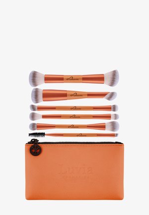 Ensemble de sept pinceaux de maquillage double embout avec des manches en or rose et des poils blancs, disposés au-dessus d'une pochette zippée orange de Luvia Cosmetics.