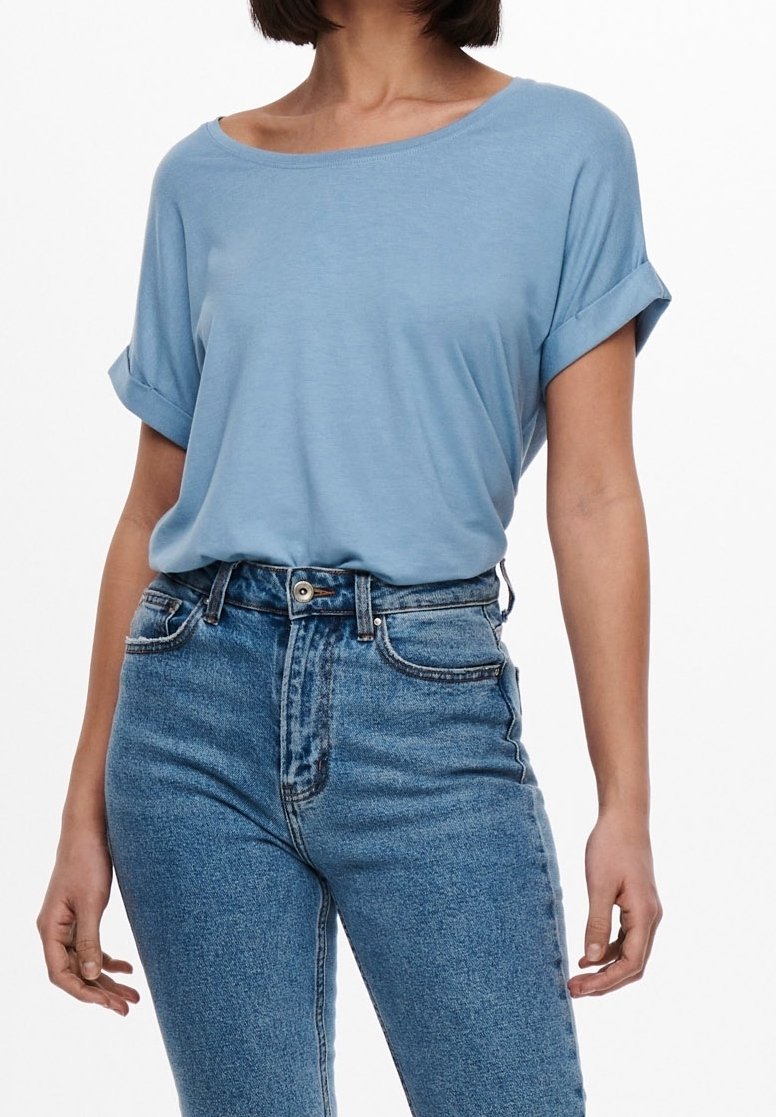 Femme portant un haut à manches courtes bleu clair rentré dans un jean denim bleu taille haute, sur fond blanc.