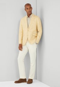 Blazer de lino amarillo claro sobre una camisa gris claro, combinado con pantalones crema y zapatos de ante marrón. Diseño simple, a medida y con corte estructurado.
