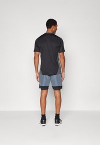 Svart sportskjorte med grå sidepaneler, kombinert med blå shorts med svart midjebånd. Modellen har på seg svarte fottøy og sokker.