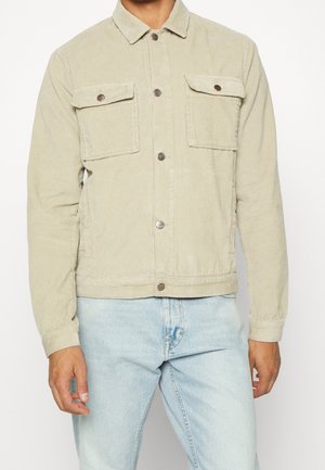 Homme portant une veste en velours côtelé beige clair avec poches poitrine boutonnées et un jean bleu clair, debout devant un fond blanc.