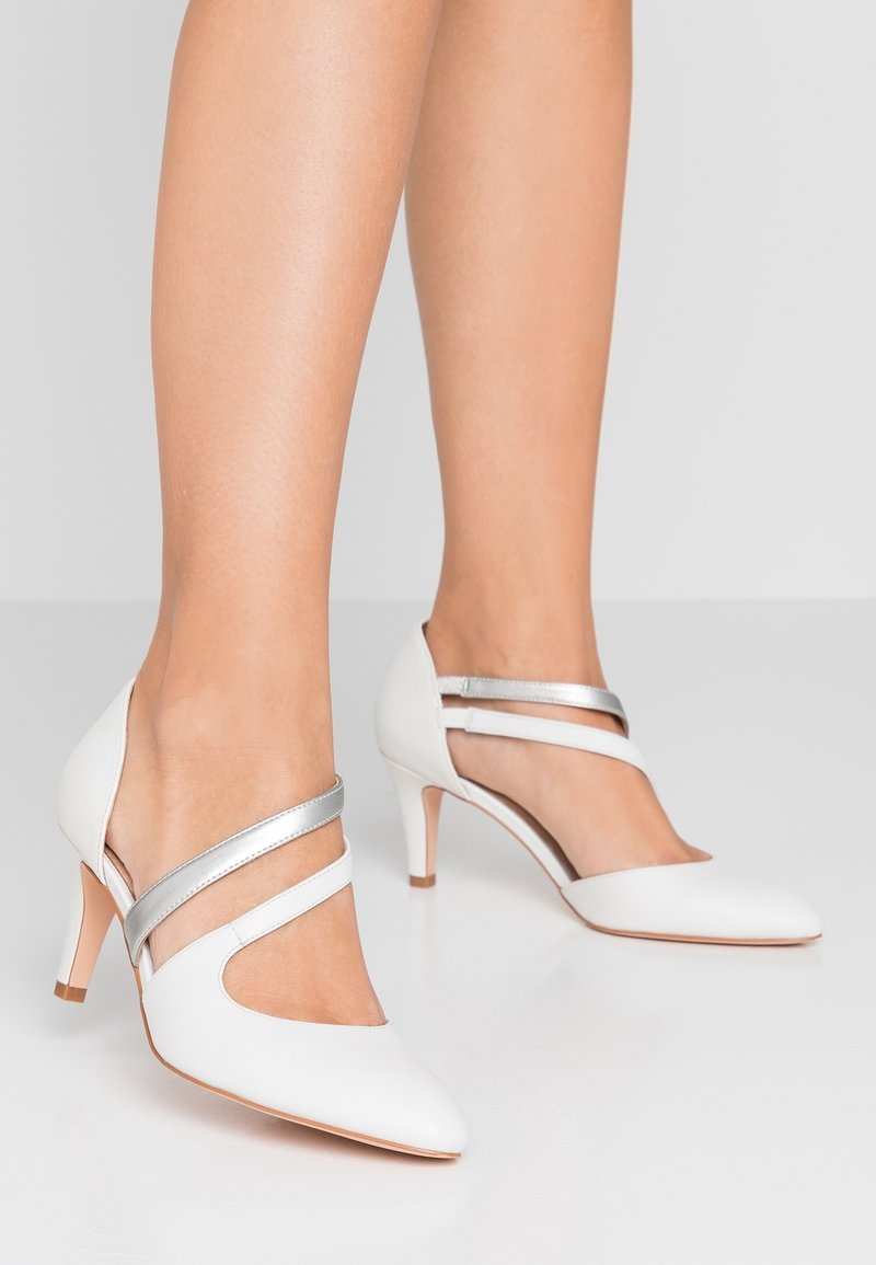 Escarpins blancs à bout pointu avec deux brides, une argentée métal et une blanche, portés pieds nus sur un fond clair uni.