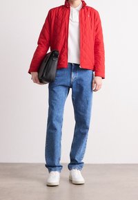 Giacca rossa trapuntata, polo bianca, jeans di denim blu e sneakers bianche. Borsa a tracolla nera tenuta nella mano sinistra. Materiali a trama.