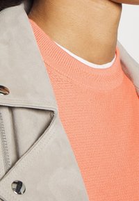 Hellgrauer Wildlederjackenkragen mit silberner Hardware über einem strukturierten, korallenfarbenen Strickpullover mit einem gerippten Ausschnitt. Weißes Unterhemd sichtbar.