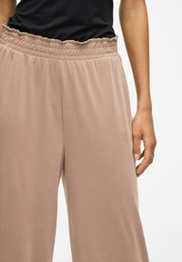 Pantalones suaves, ligeros y beiges con cintura elástica fruncida; corte holgado; textura suave; combinados con una blusa negra.