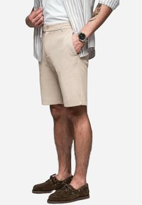 Shorts de algodón beige con bolsillos laterales, corte ajustado y dobladillo limpio. Combinados con una camiseta a rayas y zapatos náuticos de ante marrón.