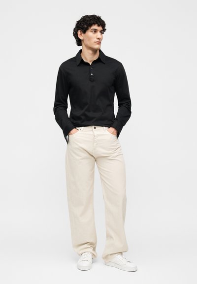 Νέος άνδρας στέκεται με τα χέρια στις τσέπες, φορώντας μαύρο πουκάμισο με μακριά μανίκια, μπεζ φαρδιά παντελόνια και λευκά αθλητικά παπούτσια μπροστά σε απλό φόντο.