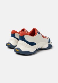 Sneakers bianchi e blu navy con una tomaia in mesh testurizzato e suede, dettagli rossi e suola in gomma Vibram. Presentano lacci decorativi e branding.