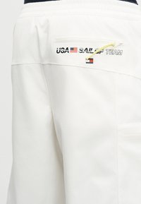 Shorts de voile blancs avec poche zippée, drapeau américain et inscription « USA SAIL GP TEAM » imprimée près de la ceinture.