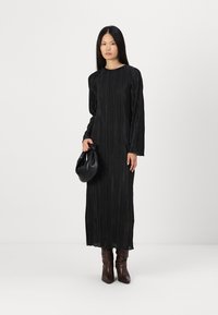 Μαύρο πλισέ φόρεμα μακρύ μανίκι με στρογγυλή λαιμόκοψη, συνδυασμένο με μαύρη τσάντα και καφέ μπότες μέχρι το γόνατο, παρουσιασμένο πάνω σε ουδέτερο φόντο.