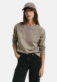 Lichte taupe sweatshirt met ronde hals en geribbelde manchetten, gedragen met hoge taille zwarte jeans. Model draagt een grijze en bruine pet met tekst.