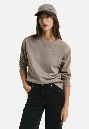 Helles taupe Sweatshirt mit Rundhalsausschnitt und gerippten Bündchen, kombiniert mit hochgeschnittenen schwarzen Jeans. Das Model trägt eine grau-braune Mütze mit Aufschrift.