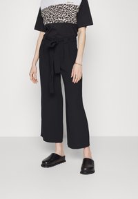 Mujer vestida con pantalones de pierna ancha negros con un cinturón atado, una blusa de manga corta negra con estampado de leopardo, y unos zapatos negros sin cordones.