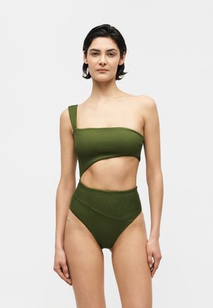 Femeie cu păr scurt și închis la culoare, purtând un bikini verde cu o singură bretea și decupaje, stând în fața unui fundal simplu și deschis la culoare.