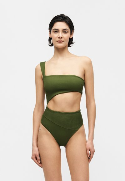 Femme aux cheveux courts et foncés portant un bikini vert à une épaule avec des découpes, debout devant un fond clair uni.