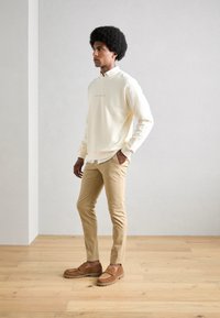 Lys beige sweatshirt, brune chinos og brune ruskind slip-on sko. Sweatshirten har et lille, præget logo. Glatte teksturer, afslappet pasform.