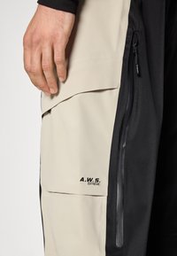 Beige a černé nepromokavé kalhoty s kapsou na zip, detailem loga "A.W.S. EXTREME" a texturovanou látkou.