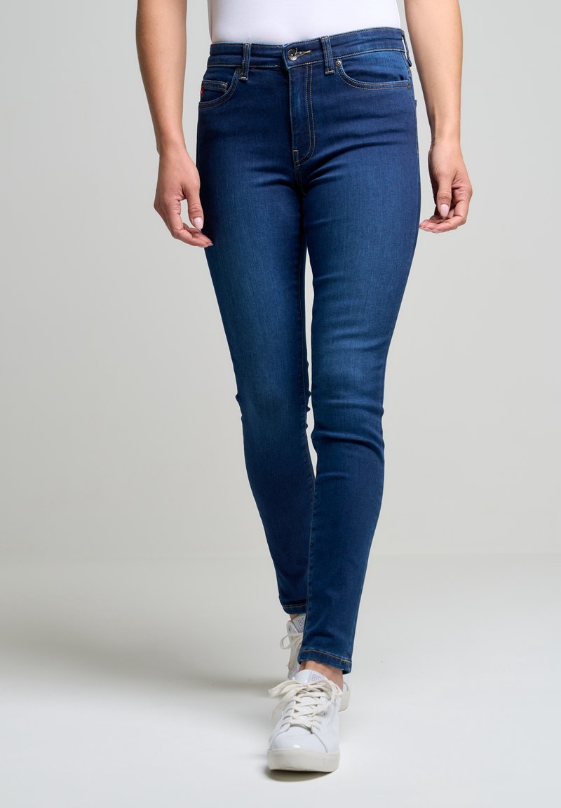 Vaqueros ajustados de mezclilla azules, con una cintura de altura media, cinco bolsillos y un cierre clásico de botón. Combinados con zapatillas blancas.