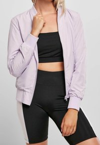 Jasnolawendowa kurtka bomber z prążkowanymi mankietami i zapięciem na zamek, noszona na czarnej bluzce crop top i czarnych spodenkach rowerowych, podkreślająca dopasowany krój.