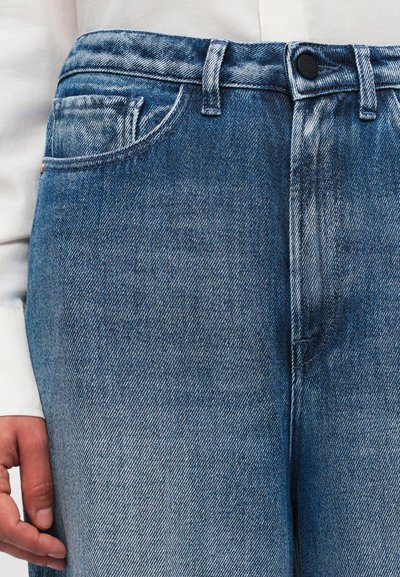 Suurendatud vaade sinistest denimipükstest, millel on kulunud tekstuur, vööpäid, nupp ja käsi, mis puhkab tasku kõrval valge särgi varruka vastu.