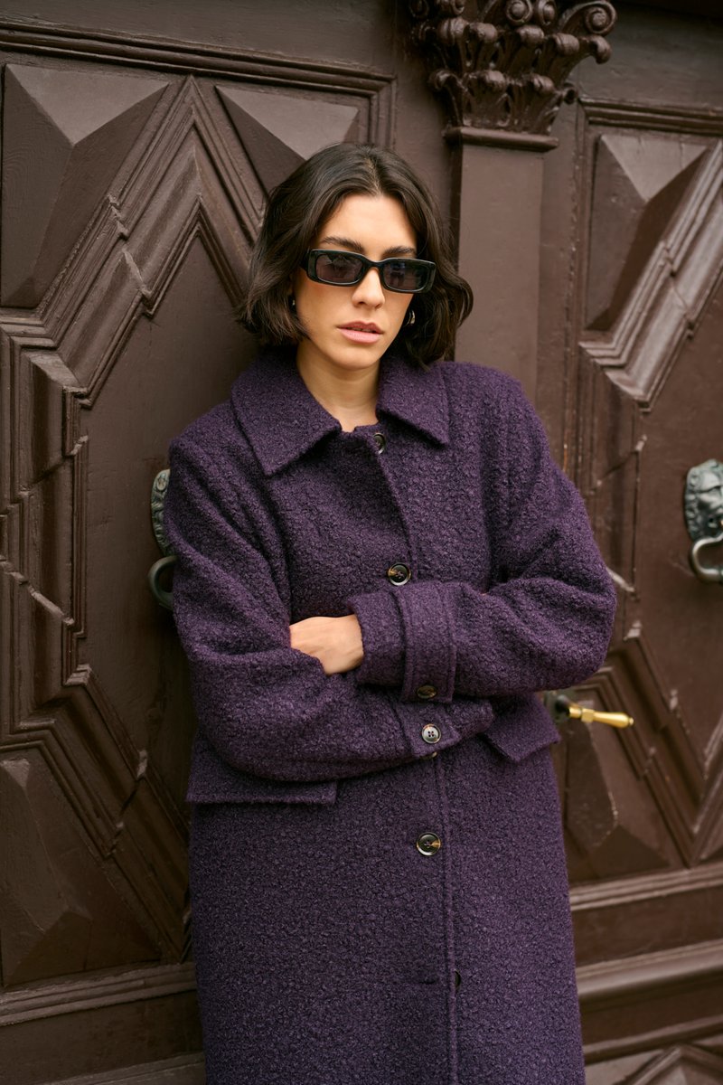 Cappotto di lana viola con tessuto testurizzato, ampio colletto e chiusura con bottoni. Modella che indossa occhiali da sole neri, in piedi contro uno sfondo di porta marrone.