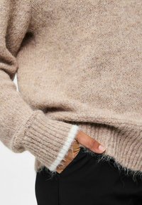 Oseba, oblečena v bež puhasto pulover s podrobnostmi v bež barvi na manšetah, ima roko skrito v žepu črnih hlač.