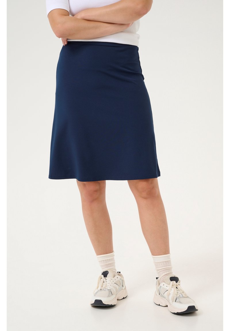 Personne portant une jupe bleu marine jusqu'aux genoux, des chaussettes blanches et des chaussures de sport blanches, debout, les bras croisés, devant un fond uni.