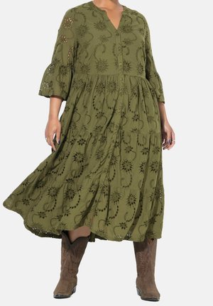 Femme portant une robe boutonnée vert olive avec des motifs floraux découpés et des bottes de cowboy marron sur un fond uni.