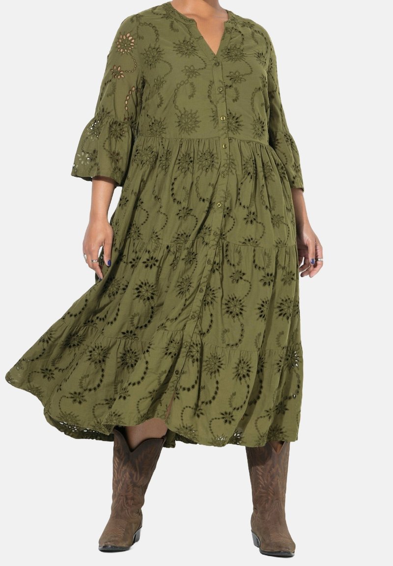 Femme portant une robe boutonnée vert olive avec des motifs floraux découpés et des bottes de cowboy marron sur un fond uni.