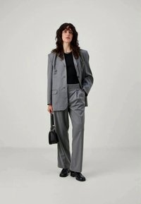 Pelēka piegriezta žakete un atbilstošas bikses no gludas auduma, apvienotas ar melnu topu. Modelis tur mazu melnu somiņu un valkā melnus apavus.