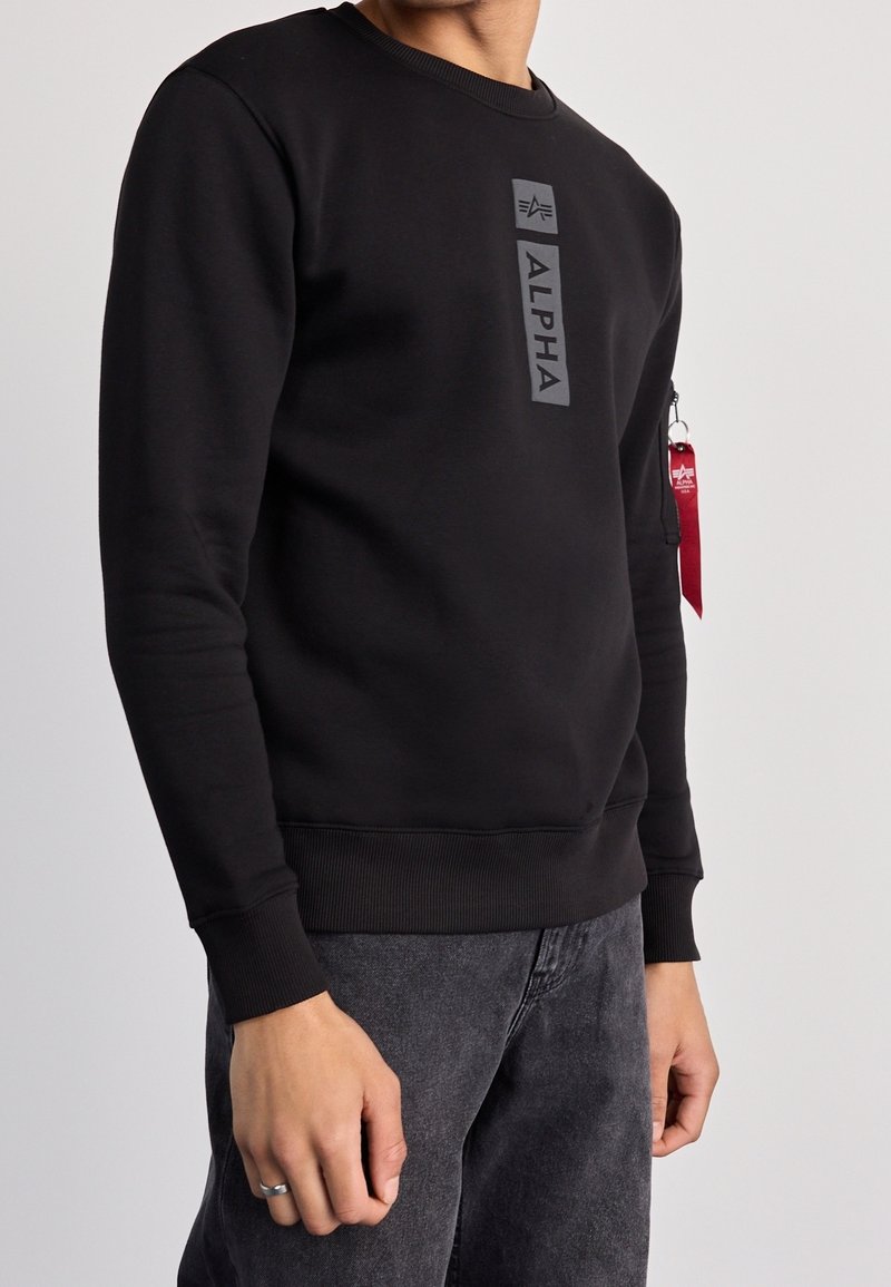 Personne portant un sweat-shirt noir avec un logo vertical "ALPHA" et une étiquette porte-clés rouge sur la poche de la manche gauche, associée à un jean gris foncé.