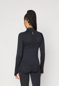 Muguras ar garajam piedurknēm, pieguļošs sporta tops melnā audumā. Tam ir augsts apkakle un teksturēts panelis aizmugurē. Iekļautas ir pirkstu caurumi pie piedurknēm.