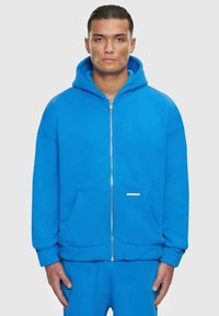 Blaues Zip-Hoodie mit großer Kapuze, Fronttaschen und gerippten Bündchen. Glatter Stoff und minimalistisches Design mit einem kleinen Etikett auf der Vorderseite.