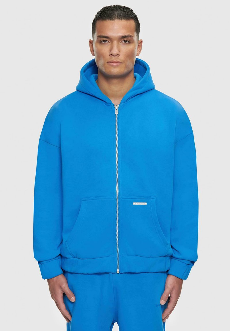 Blaues Zip-Hoodie mit großer Kapuze, Fronttaschen und gerippten Bündchen. Glatter Stoff und minimalistisches Design mit einem kleinen Etikett auf der Vorderseite.