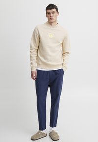 Sudadera beige de cuello redondo con estampado de logotipo, combinada con pantalones azul marino y zapatos tipo slip-on beige. El modelo se encuentra de pie frente a un fondo neutro.